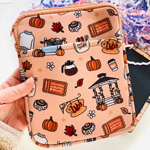 May include: A peach tablet sleeve with a zip closure, patterned with pumpkins, leaves, coffee cups, and the text "Stars Hollow Autumn Festival." The design includes a gazebo and a sign that says "Luke's."