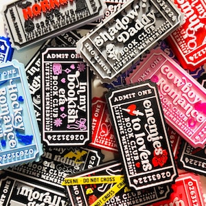 Op de afbeelding: Een verzameling kleurrijke, rechthoekige magneten in de vorm van tickets, met verschillende tekst- en grafische ontwerpen. De magneten bevatten zinnen als "Horror", "Shadow Daddy" en "Enemies to Lovers". Het kleurenpalet omvat rood, zwart, wit, roze en blauw.