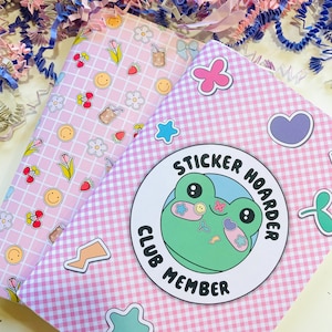 May include: Two pink notebooks with decorative patterns and stickers. One notebook has a grid pattern with cherries, flowers, and smiley faces. The other has a gingham pattern with a frog sticker that says "Sticker Hoarder Club Member".