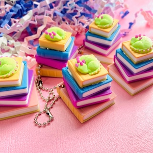 May include: A collection of miniature book charms, each with a sleeping frog design. The books are in shades of pink, blue, yellow, and purple. Some charms have a silver chain. The background is pink with colourful paper confetti.