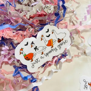 May include: A white sticker with the phrase "just a Spooky goose" and illustrations of geese in Halloween costumes. The geese are wearing a witch hat, an orange pumpkin costume, and a pumpkin. The sticker is surrounded by pink, white, and blue paper shreds.