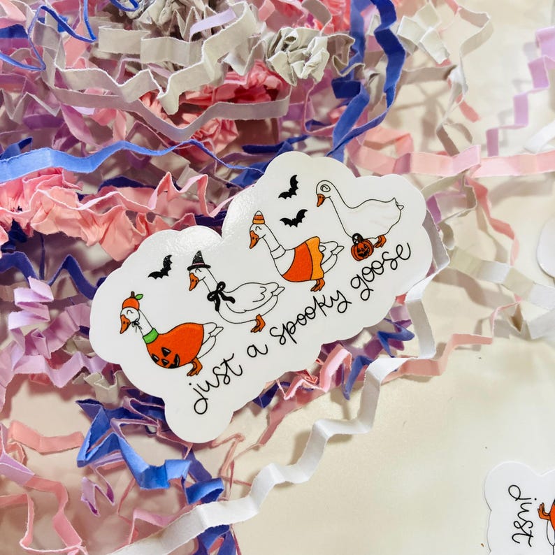 May include: A white sticker with the phrase "just a Spooky goose" and illustrations of geese in Halloween costumes. The geese are wearing a witch hat, an orange pumpkin costume, and a pumpkin. The sticker is surrounded by pink, white, and blue paper shreds.