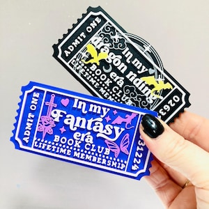 Op de afbeelding: Twee decoratieve tickets. E&eacute;n is zwart met de tekst "In my dragon riding era BOOK CLUB", de andere is blauw met "In my Fantasy era BOOK CLUB LIFETIME MEMBERSHIP". Beide hebben witte en gele accenten.
