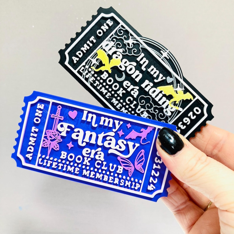 Op de afbeelding: Twee decoratieve tickets. E&eacute;n is zwart met de tekst "In my dragon riding era BOOK CLUB", de andere is blauw met "In my Fantasy era BOOK CLUB LIFETIME MEMBERSHIP". Beide hebben witte en gele accenten.