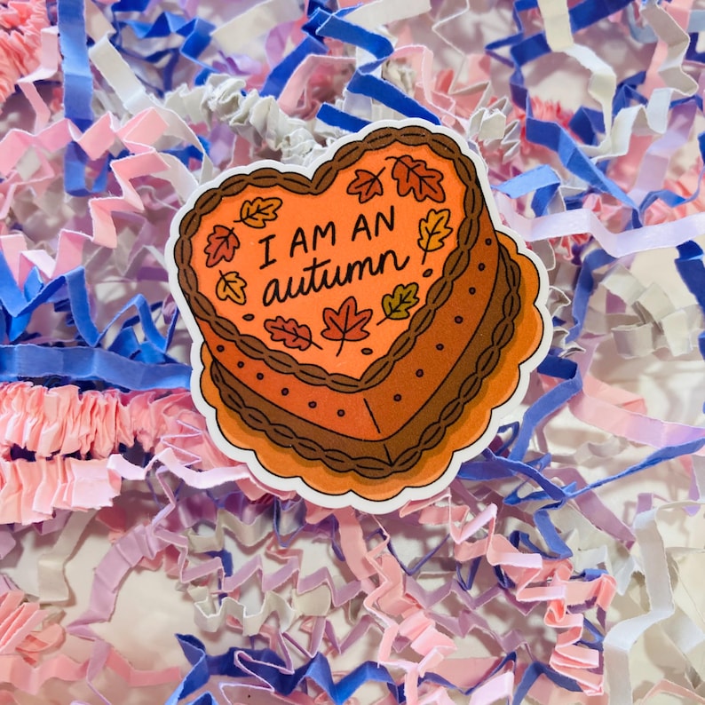 Op de afbeelding: Een hartvormige sticker met de tekst "I AM AN autumn" in zwart schrift. De sticker heeft een cake-ontwerp met oranje glazuur, bruine randen en herfstbladeren. De achtergrond is gevuld met roze, blauwe en witte papiersnippers.
