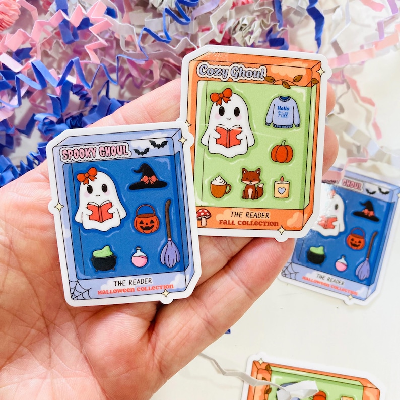 May include: A hand holds several stickers featuring a cartoon ghost reading a book. The stickers have a Halloween theme with pumpkins, witch hats, and the text "Spooky Ghoul" and "Cozy Ghoul". The stickers are in a box-like design.
