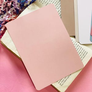May include: A pale pink rectangular card is placed on top of an open book with aged pages. The card has rounded corners and a smooth surface. The background includes other books and pink paper.
