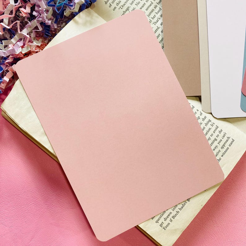 May include: A pale pink rectangular card is placed on top of an open book with aged pages. The card has rounded corners and a smooth surface. The background includes other books and pink paper.