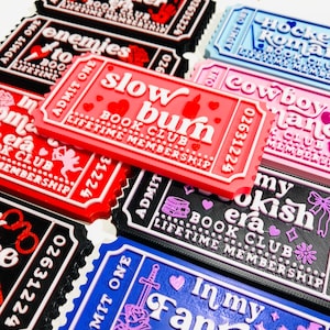 Op de afbeelding: Een verzameling ticketvormige magneten met verschillende boeken-thema's. De magneten tonen teksten als "Slow Burn Book Club" en "In My Fantasy Era". Kleuren zijn rood, zwart, blauw en roze.