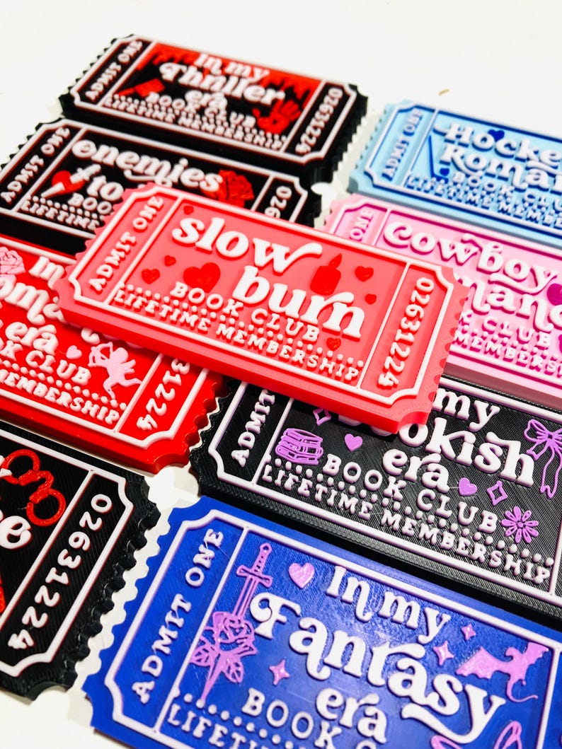 Op de afbeelding: Een verzameling ticketvormige magneten met verschillende boeken-thema's. De magneten tonen teksten als "Slow Burn Book Club" en "In My Fantasy Era". Kleuren zijn rood, zwart, blauw en roze.