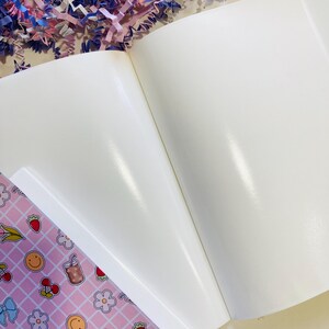 May include: Open white blank notebook with glossy pages. A pink patterned page with illustrations of cherries, strawberries, flowers, and a smiley face is visible. Decorative paper shreds in pink, blue, and purple are in the background.