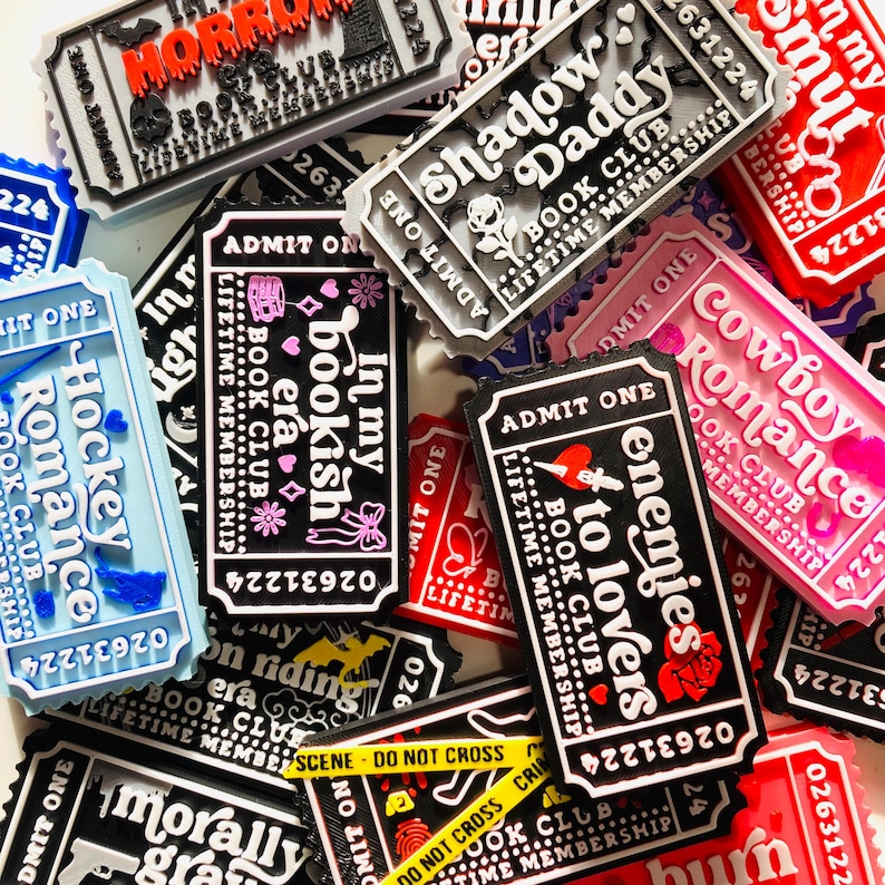 Op de afbeelding: Een verzameling kleurrijke, rechthoekige magneten in de vorm van tickets, met verschillende tekst- en grafische ontwerpen. De magneten bevatten zinnen als "Horror", "Shadow Daddy" en "Enemies to Lovers". Het kleurenpalet omvat rood, zwart, wit, roze en blauw.