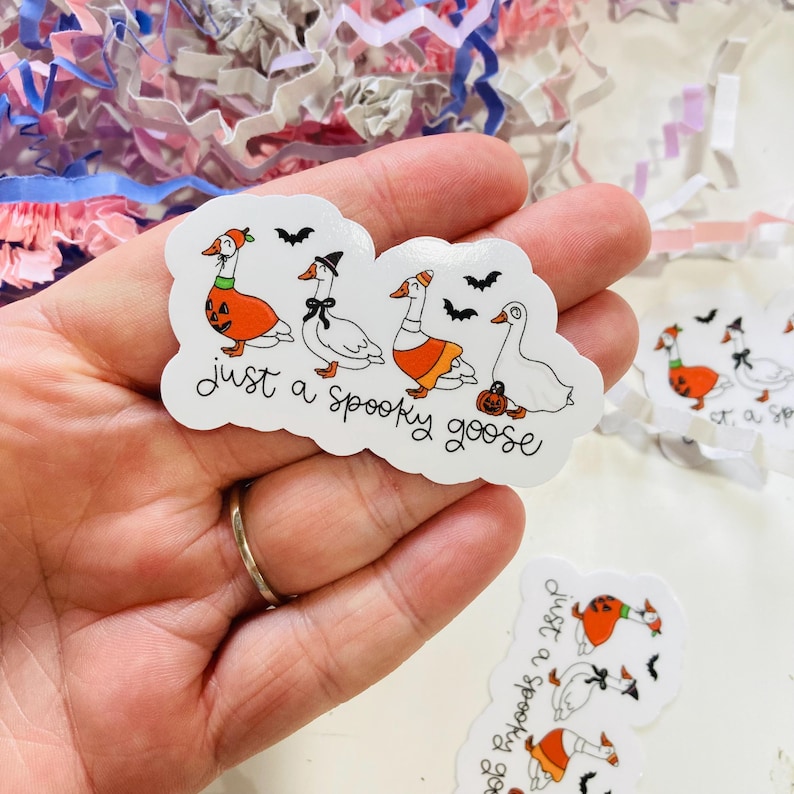 May include: A white sticker with the text "just a spooky goose" and illustrations of geese in Halloween costumes. The geese are wearing a pumpkin, a witch hat, and an orange skirt. Black bats are also included in the design.
