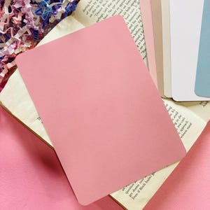 May include: A stack of blank, rounded-corner pink cards, resting on an open book with aged pages. The cards are a solid, soft pink color. Other cards in shades of white, beige, and blue are visible in the background.