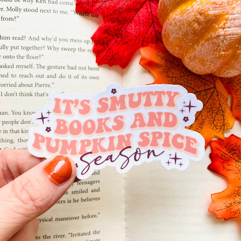 以下が含まれることがあります： ピンクと紫色の「IT'S SMUTTY BOOKS AND PUMPKIN SPICE SEASON」という文字が入ったステッカー。ステッカーは手で持たれており、背景にはカボチャと紅葉があります。