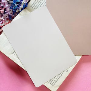 May include: A blank, off-white rectangular card, slightly angled, resting on a book with printed text. The card is positioned over a pink surface, with a hint of a tan surface visible in the background.