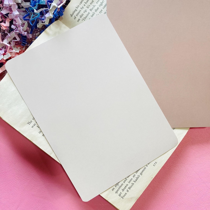 May include: A blank, off-white rectangular card, slightly angled, resting on a book with printed text. The card is positioned over a pink surface, with a hint of a tan surface visible in the background.