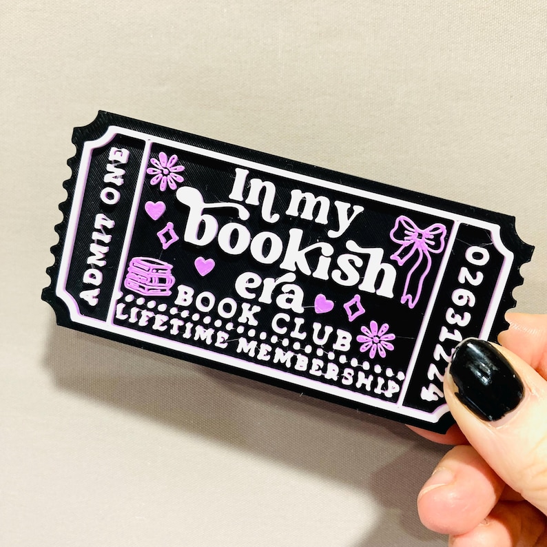 Op de afbeelding: Een zwart en roze ticketvormig item met de tekst "In my bookish era" en "Book Club Lifetime Membership". Het ticket heeft een decoratieve strik, hartjes en bloemmotieven.