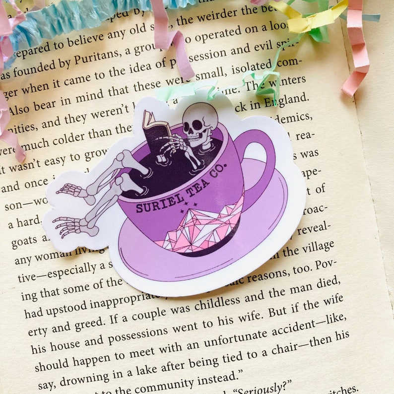 May include: A purple teacup with a skeleton sitting inside, reading a book. The teacup has a pink and white crystal design and the text "SURIEL TEA CO." on the side.
