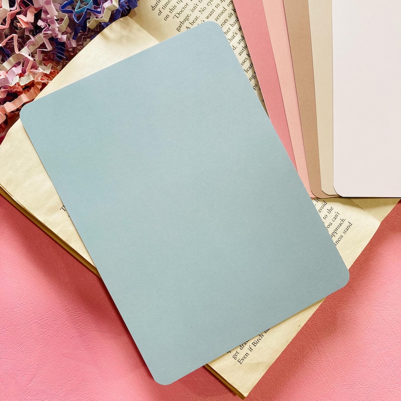 May include: A stack of pastel-colored cardstock paper in shades of blue, pink, beige, and white. A light blue card is on top of an open book with aged pages. The background is a pink surface.