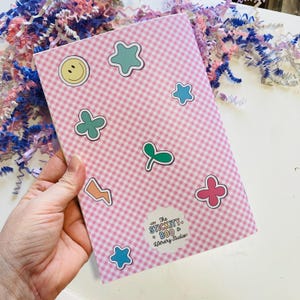 May include: A pink and white checkered notebook adorned with colorful stickers in various shapes, including stars, flowers, and a smiley face. The notebook has a sticker that says "The Stickity Boo & Literary Studios".