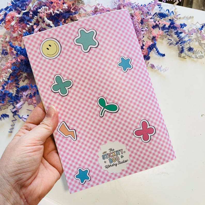 May include: A pink and white checkered notebook adorned with colorful stickers in various shapes, including stars, flowers, and a smiley face. The notebook has a sticker that says "The Stickity Boo & Literary Studios".