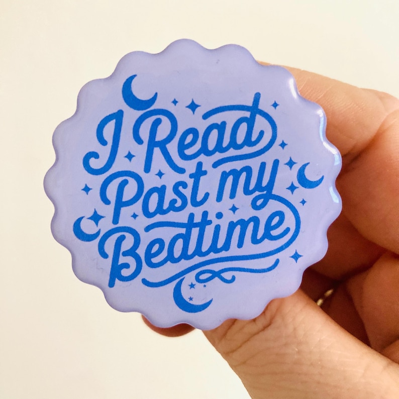 May include: A light purple, scalloped-edge button with the text "I Read Past my Bedtime" in blue script. The design includes crescent moons and stars. A hand is holding the button.