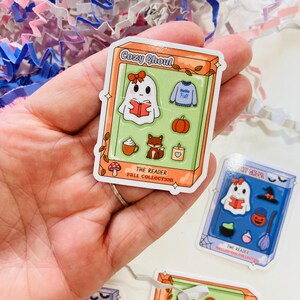 May include: A sticker featuring a cartoon ghost reading a book, with a bow, inside a box labeled "Cozy Ghoul". Other images include a sweater, pumpkin, fox, candle, and coffee cup. The text "The Reader Fall Collection" is also present.