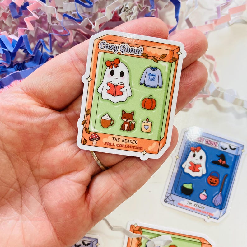 May include: A sticker featuring a cartoon ghost reading a book, with a bow, inside a box labeled "Cozy Ghoul". Other images include a sweater, pumpkin, fox, candle, and coffee cup. The text "The Reader Fall Collection" is also present.