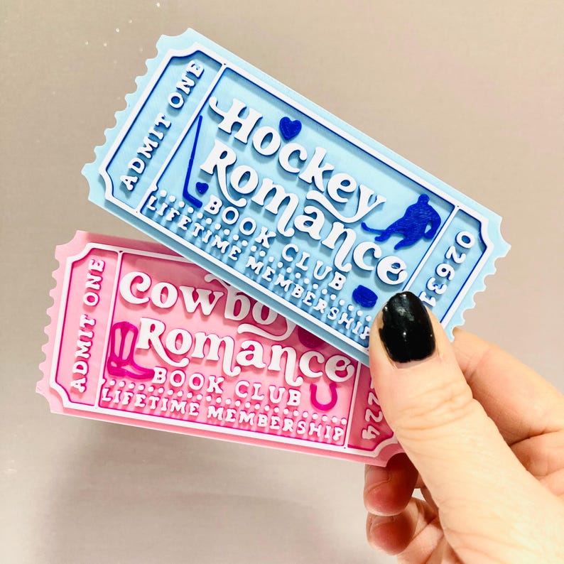 Op de afbeelding: Twee decoratieve magneten in de vorm van een ticket. E&eacute;n is lichtblauw met de tekst "Hockey Romance Book Club" en een silhouet van een hockeyspeler. De andere is roze met de tekst "Cowboy Romance Book Club" en een cowboylaarsafbeelding. Beide luiden "Lifetime Membership".