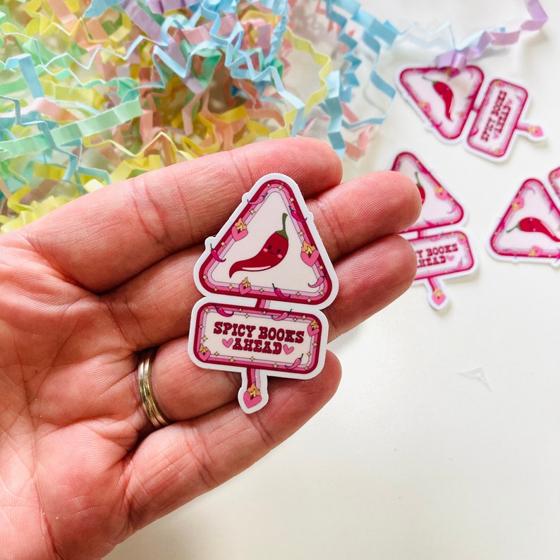 May include: A pink and white sticker with a red chilli pepper and the text "Spicy Books Ahead" with hearts. The sticker is shaped like a warning sign.