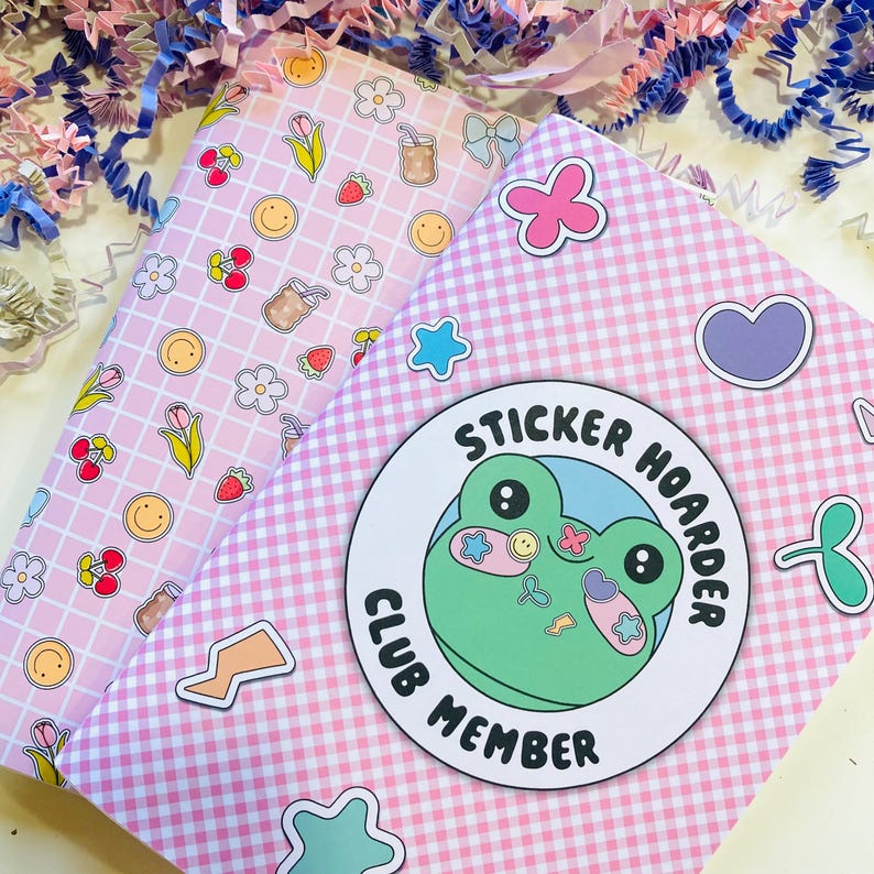 May include: Two pink notebooks with decorative patterns and stickers. One notebook has a grid pattern with cherries, flowers, and smiley faces. The other has a gingham pattern with a frog sticker that says "Sticker Hoarder Club Member".