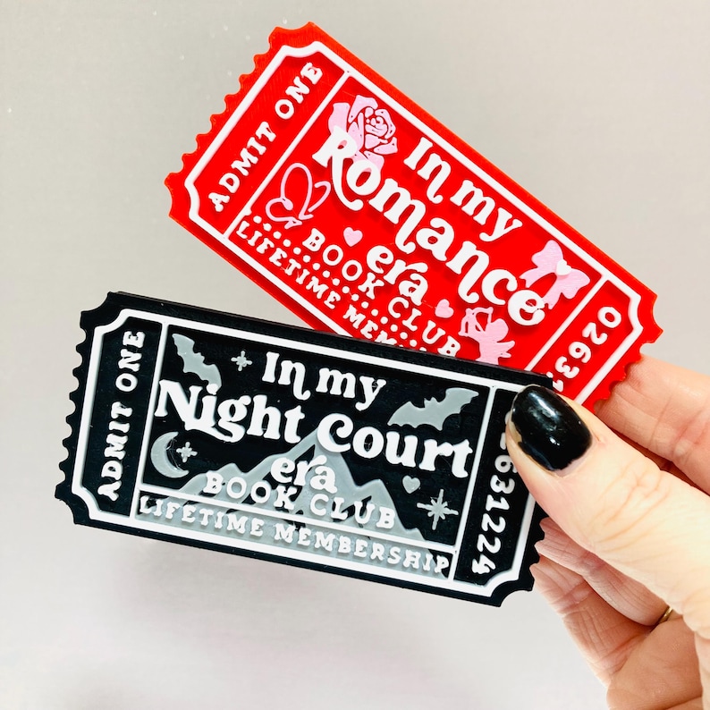 Op de afbeelding: Twee decoratieve items in de vorm van een ticket. E&eacute;n is rood met witte tekst die luidt "In my Romance era BOOK CLUB" en "ADMIT ONE". De andere is zwart met witte tekst die luidt "In my Night Court era BOOK CLUB" en "ADMIT ONE".