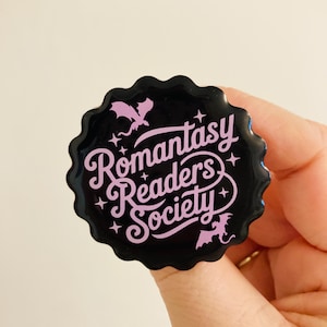 May include: A black, scalloped-edge button with the text "Romantasy Readers Society" in pink script. The button features two pink dragon silhouettes and star accents, perfect for book lovers.