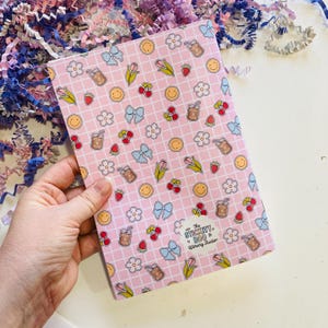May include: A pink notebook with a grid pattern and repeating illustrations of bows, flowers, cherries, strawberries, smiley faces, and iced drinks. The notebook is held in a hand and has the text "The Stickity Boo Literary Studio".