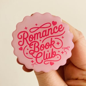 May include: A pink, scalloped-edge button with the words "Romance Book Club" in a cursive font. The text is in a darker pink, with hearts and stars as decorative elements. The button is held in a hand.