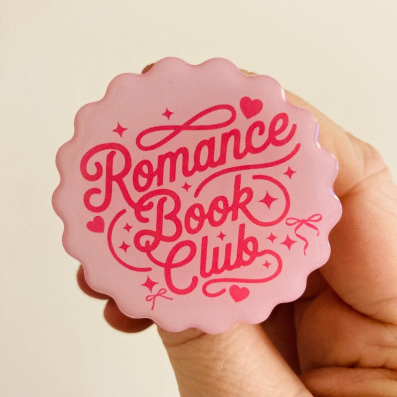 May include: A pink, scalloped-edge button with the words "Romance Book Club" in a cursive font. The text is in a darker pink, with hearts and stars as decorative elements. The button is held in a hand.