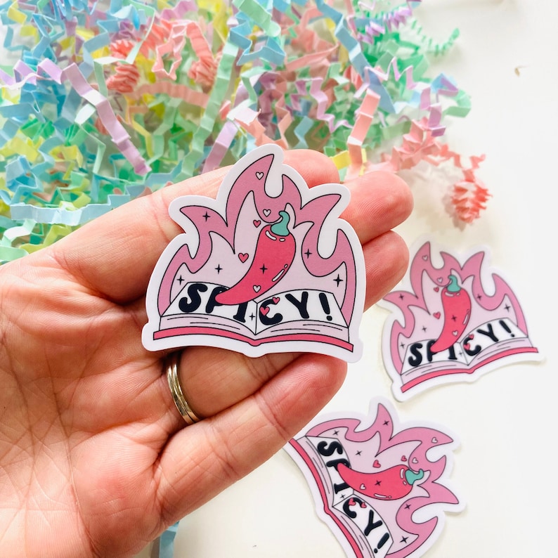 Op de afbeelding: Een roze en witte sticker met een rode chilipeper op een boek met de tekst "SPICY!" in roze letters. De chilipeper staat in brand met roze vlammen.