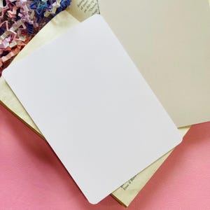 May include: A blank white card with rounded corners, resting on a stack of papers. The card is angled, revealing a glimpse of the underlying pages, including a beige page and a page with text. The background includes pink and floral elements.