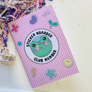 May include: A pink and white checkered notebook with various sticker designs. The notebook features a large circular sticker with a green frog and the text "STICKER HOARDER CLUB MEMBER". Additional stickers include a heart, flower, star, and lightning bolt.