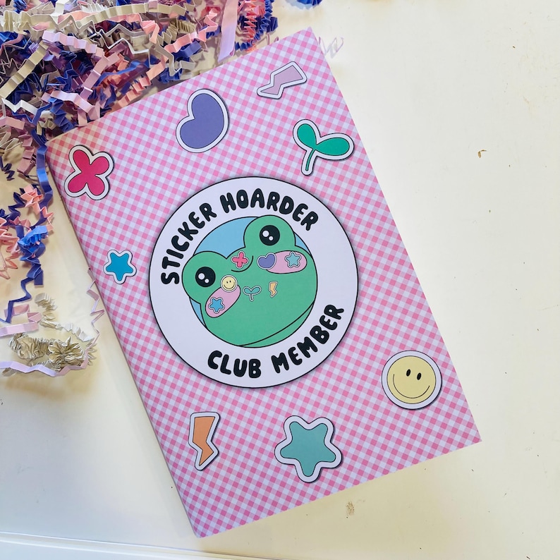 May include: A pink and white checkered notebook with various sticker designs. The notebook features a large circular sticker with a green frog and the text "STICKER HOARDER CLUB MEMBER". Additional stickers include a heart, flower, star, and lightning bolt.