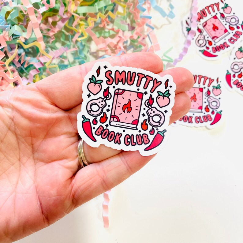 May include: A white sticker with the text "SMUTTY BOOK CLUB" in pink and black lettering. The sticker features a pink book with a flame, handcuffs, chili peppers, and peaches.