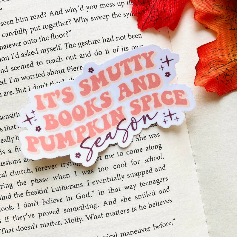 以下が含まれることがあります： ピンクとピーチの色合いで「IT'S SMUTTY BOOKS AND PUMPKIN SPICE Season」というテキストが入ったダイカットステッカー。ステッカーは白い縁取りがあり、小さな花のアクセントとスターバーストで装飾されています。ステッカーは開いた本の上に置かれています。