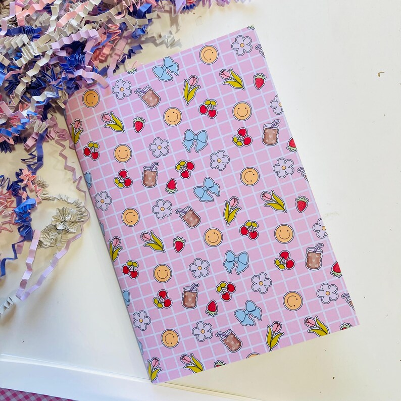May include: A pink notebook with a grid pattern and repeating illustrations of bows, flowers, cherries, strawberries, smiley faces, and drinks. The notebook is surrounded by colorful paper shreds.