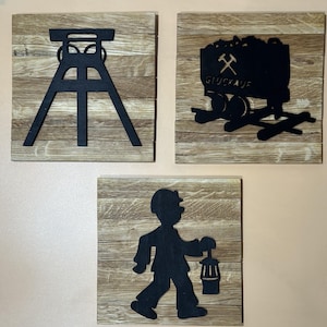 Könnte beinhalten: Drei Holz-Wandplaketten mit schwarzen Silhouetten-Designs, die ein Bergbau-Thema darstellen. Die erste Plakette zeigt ein Fördergerüst, die zweite zeigt einen Grubenwagen mit dem Text "Glückauf" und die dritte zeigt einen Bergmann mit einer Laterne.