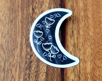 May include: A black and white sticker of a crescent moon with white floral designs inside.