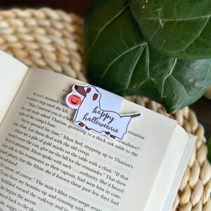 May include: A white dachshund with a brown nose and tail holds a small orange pumpkin with a jack-o-lantern face. The bookmark is white with the text "happy halloweenie" in black cursive writing.