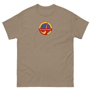May include: A tan t-shirt featuring a circular graphic with the text "ИНТЕРКОСМОС" in red letters. The graphic includes a yellow and red rocket launching from a blue circle, set against a red border. The shirt is made of a soft material.