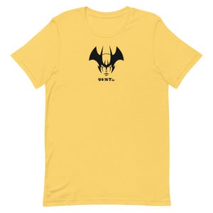 May include: A yellow t-shirt featuring a black graphic of a demon-like face with horns and the Japanese text "デビルマン" below it. The design is centered on the front of the short-sleeved shirt.