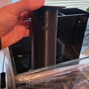 Puede incluir: Un filtro de acuario cilíndrico negro con el texto "My Reef" se sostiene frente a un acuario negro. El filtro es de plástico y tiene una parte superior ensanchada. El acuario está envuelto en plástico transparente.